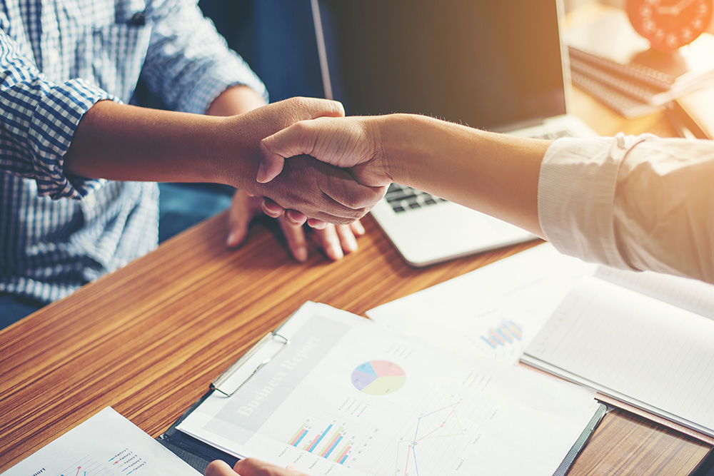 Two people shaking hands across a table with business charts and a laptop.